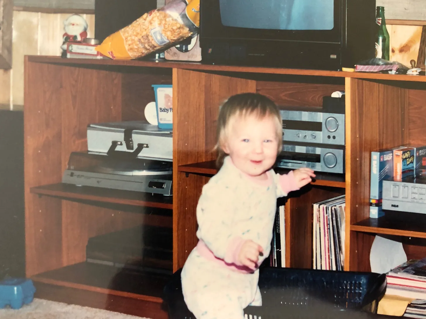 A toddler in pajamas, mid-laugh, standing by a wood-paneled stereo cabinet.